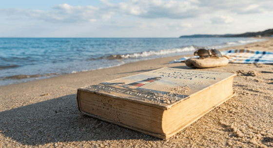 A worn book with a lighthouse illustration lying on sandy beach with sunglasses on a rock beside it